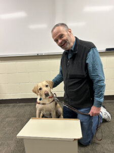 Jack kneels next to yellow guide dog Juliet in a classroom setting 