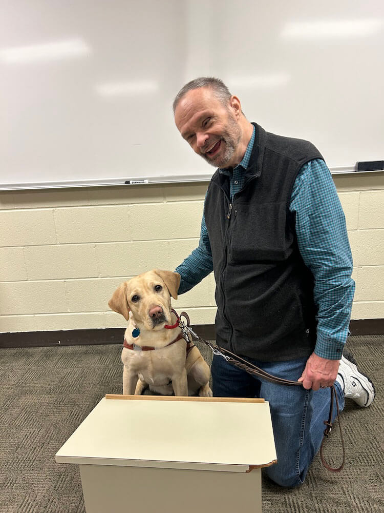 Jack kneels next to yellow guide dog Juliet in a classroom setting