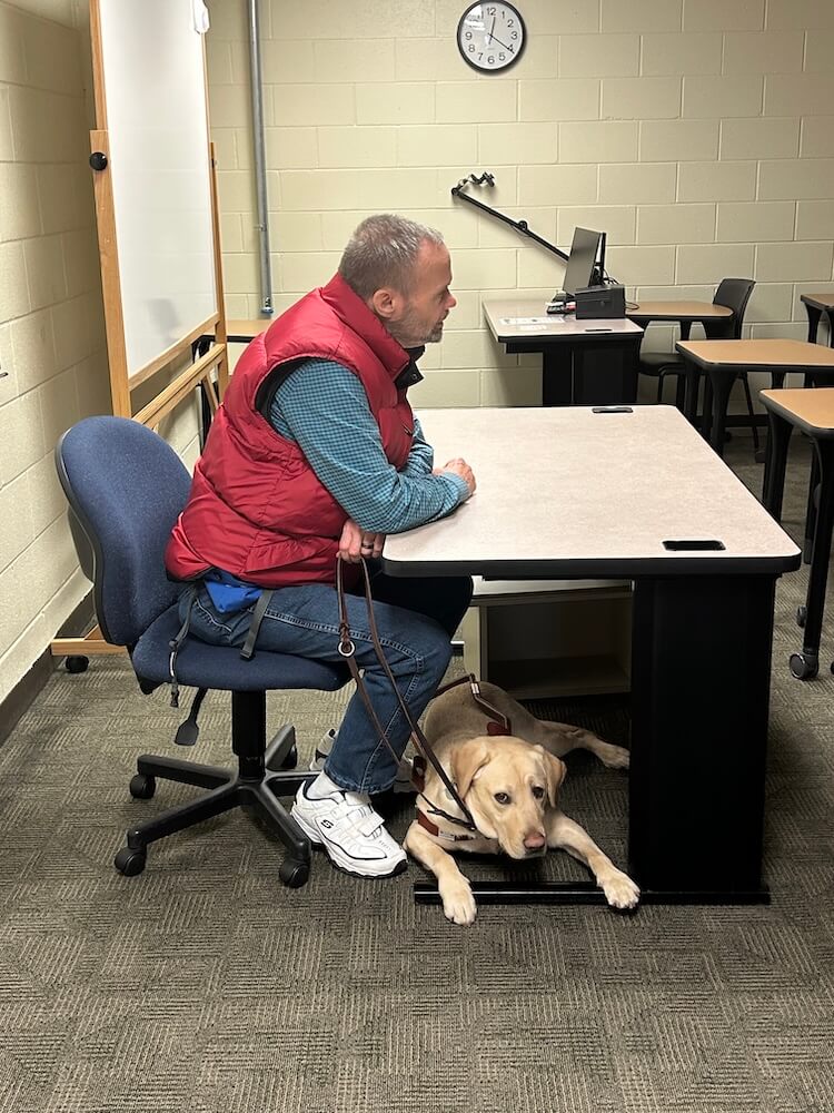 Jack sits at a desk at the front of a classroom as Juliet rests at his feet