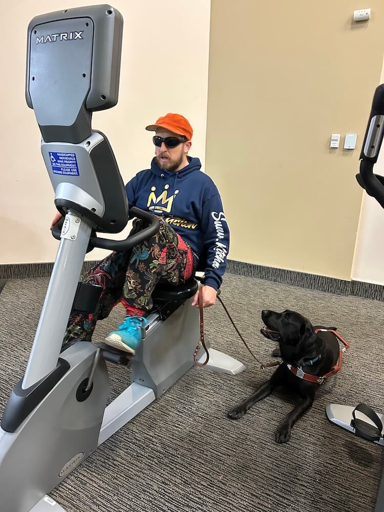 Yodel lies on the floor looking towards Jason who pedals on a large exercise bike