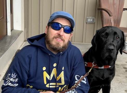 At a residential entrance, Jason squats down next to black Lab guide dog Yodel as they look to the camera.
