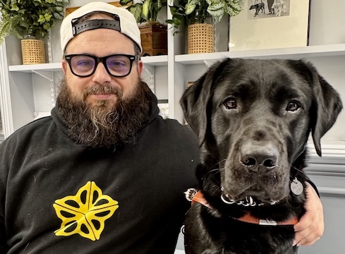 Justin sits next to black Lab guide dog Expo for their team portrait in front of shelves filled with various items