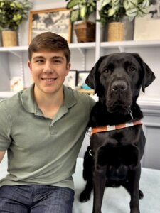 Lucien and black Lab guide dog Garrett sit in front of shelves filled with various items for their team portrait