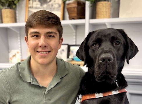 Lucien and black Lab guide dog Garrett sit in front of shelves filled with various items for their team portrait