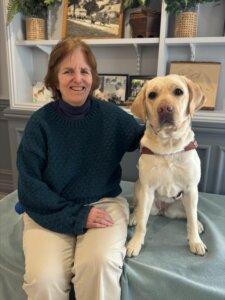 Martha and black Lab guide dog Quinnah sit in front of shelves filled with various items for their team portrait