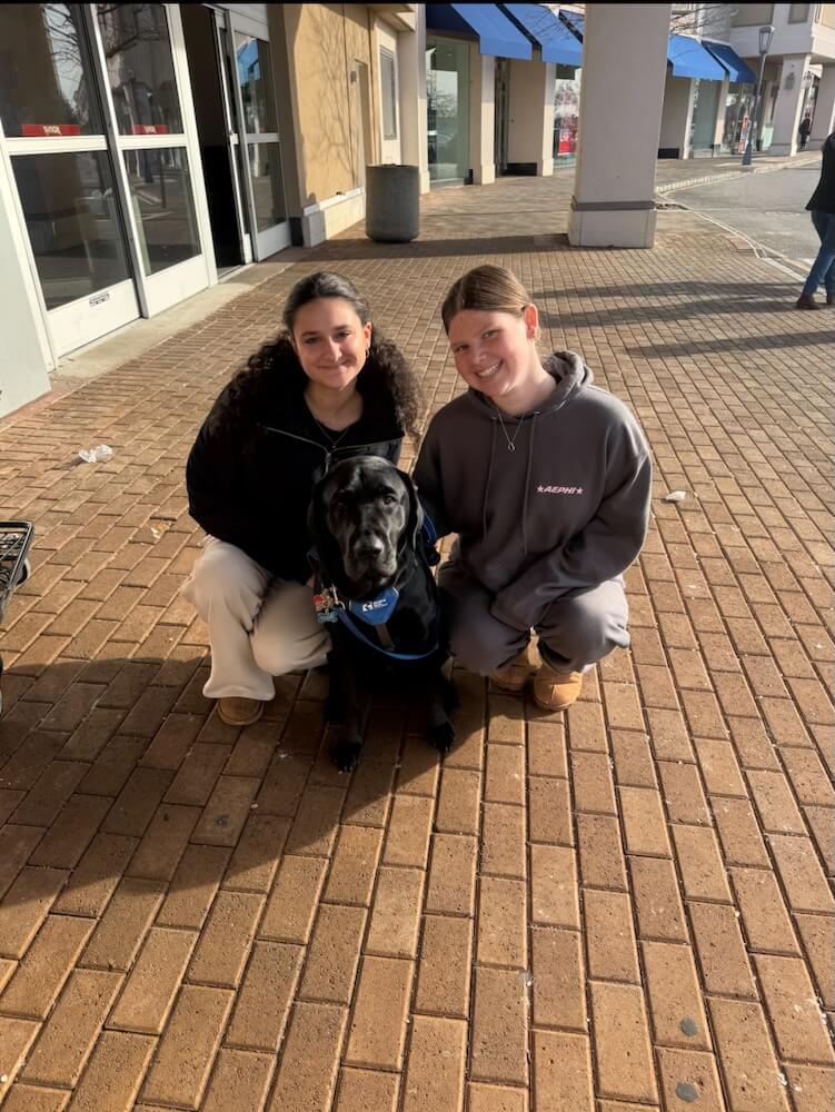 Pup Yodel poses with his raisers, who kneel next to him on a brick walkway in a shopping area
