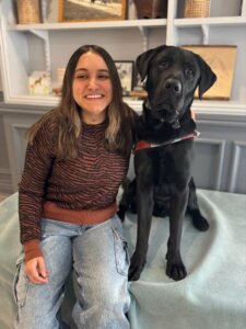 Sofia and black Lab guide dog Quinlan sit in front of shelves filled with various items for their team portrait