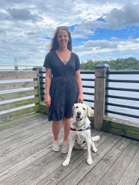 Abigail standing on a wooden deck with guide dog Kit, a yellow Labrador retriever wearing a harness, beside a river with railing, trees, and water visible in the background.