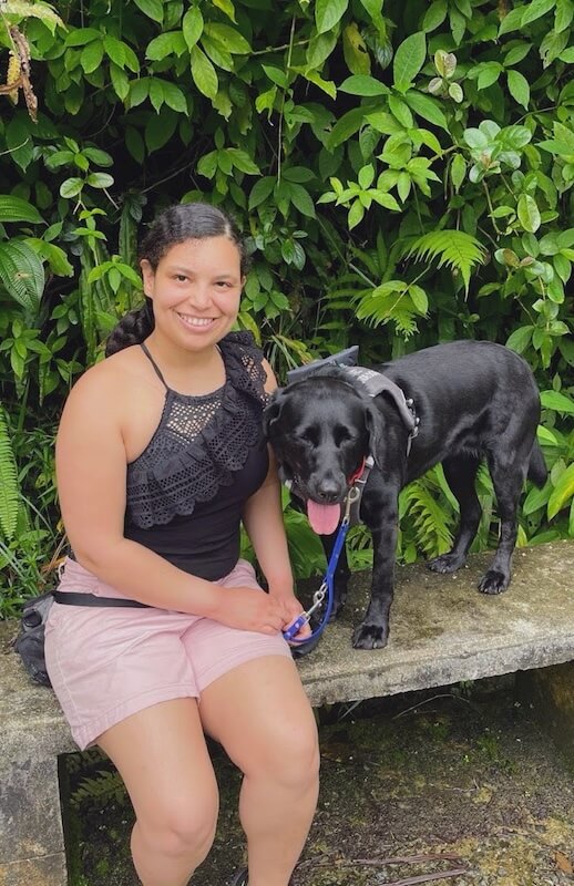 Christina seated on a stone bench outdoors with guide dog Arcadia, a black Labrador retriever wearing a harness, surrounded by green foliage.