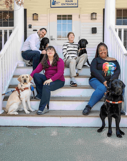 Four students from a March 2026 class sit on the front steps of the Training School main entrance with their guide dogs on a warm day during class.
