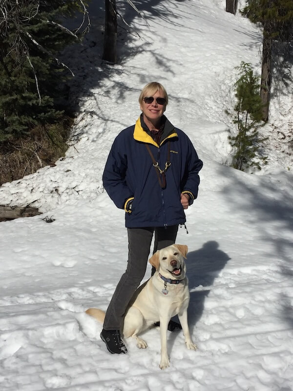 Deni standing beside yellow Lab guide dog Alberta close together in a snowy Montana location near evergreens.