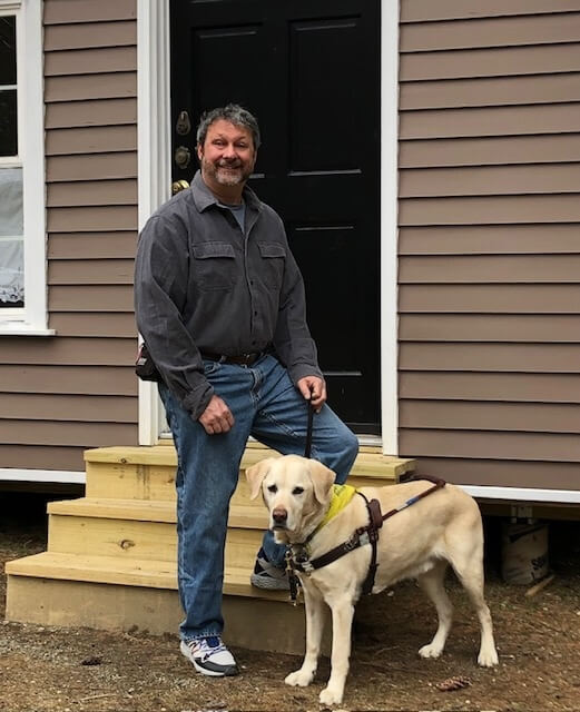 Eric standing outside a house with guide dog Ryan, a yellow Labrador retriever wearing a harness, positioned on wooden steps near a front door.