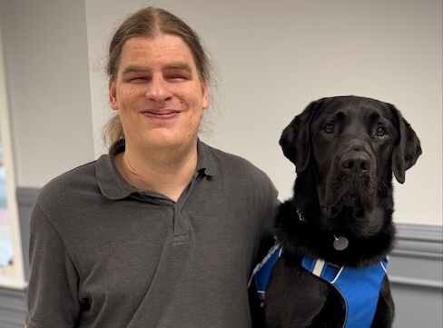 Jim and black Lab guide dog Orlando sit for their team portrait in front of shelves filled with various items