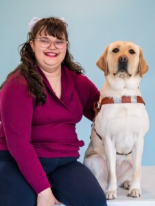 Kaitlynn and yellow Lab guide dog Raquel, sit together against a pale blue background for their team portrait