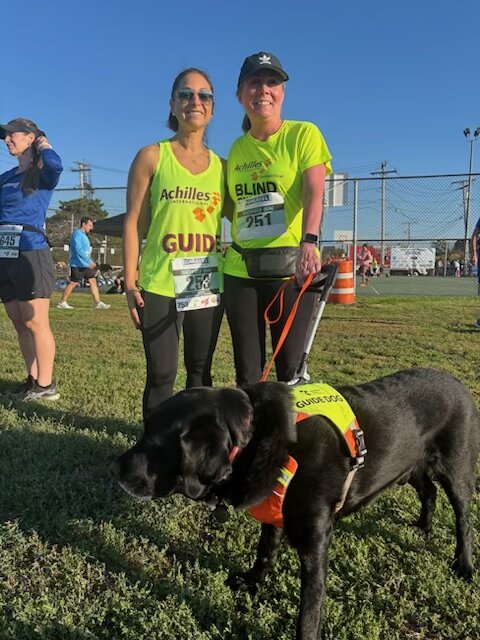 Karen holds the handle of Flint's harness as she stands with her sighted guide, both in