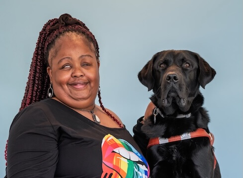Nicole and black Lab guide dog Everley, sit together against a pale blue background for their team portrait
