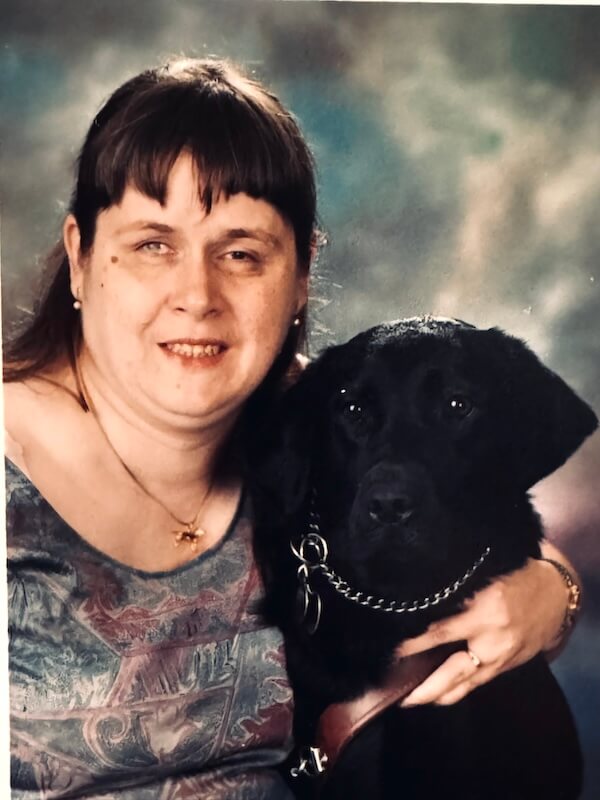 **Alt text:** “Rita with guide dog Garland, a black dog wearing a collar and harness, seated together against a studio-style background.”