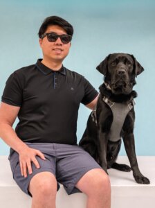 Benjamin and black Lab guide dog Owen pose for their graduate team portrait against a blue background.