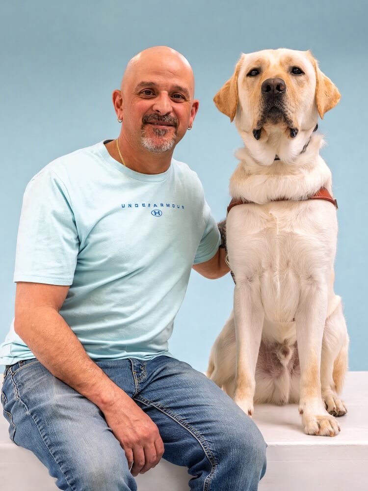 Brian and yellow Lab guide dog Moose pose for their graduate team portrait against a blue background.