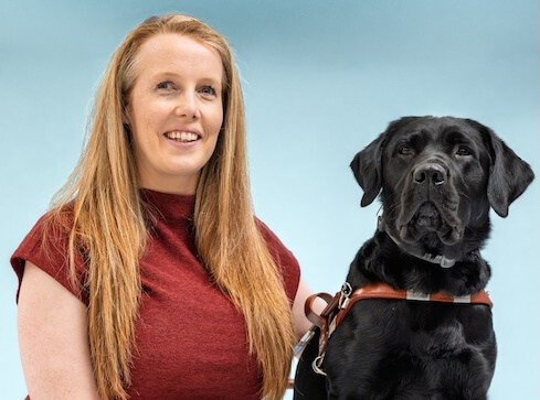 Christine and black Lab guide dog Vader pose for their graduate team portrait against a blue background.