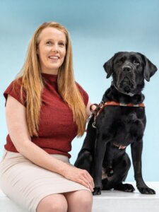 Christine and black Lab guide dog Vader pose for their graduate team portrait against a blue background.