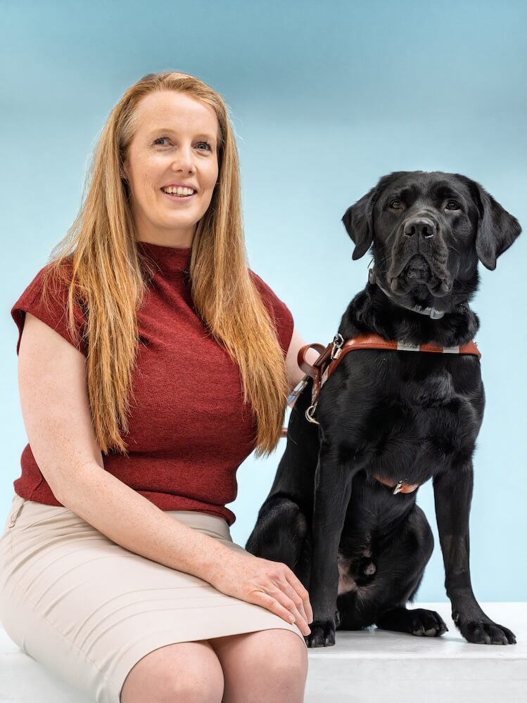 Christine and black Lab guide dog Vader pose for their graduate team portrait against a blue background.