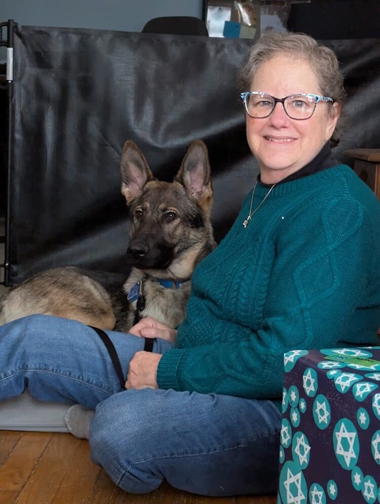 GS pup Rhiana lies on a dog bed with ears alertly up as raiser Marci sits on the floor with her