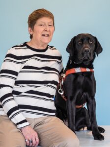 Kathy and black Lab guide dog Avis pose for their graduate team portrait against a blue background.