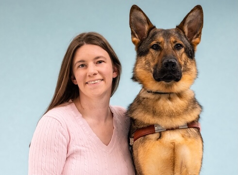 Meagan and German Shepherd Guide Dog Rhiana pose for their graduate portrait in front of a light blue background