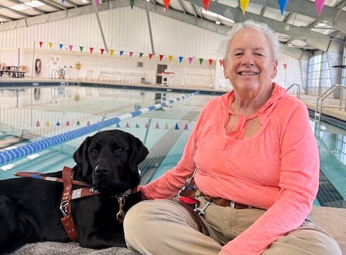 Black Lab guide dog Robbie lies next to her handler Oleta who sits on the paved rim of an indoor pool
