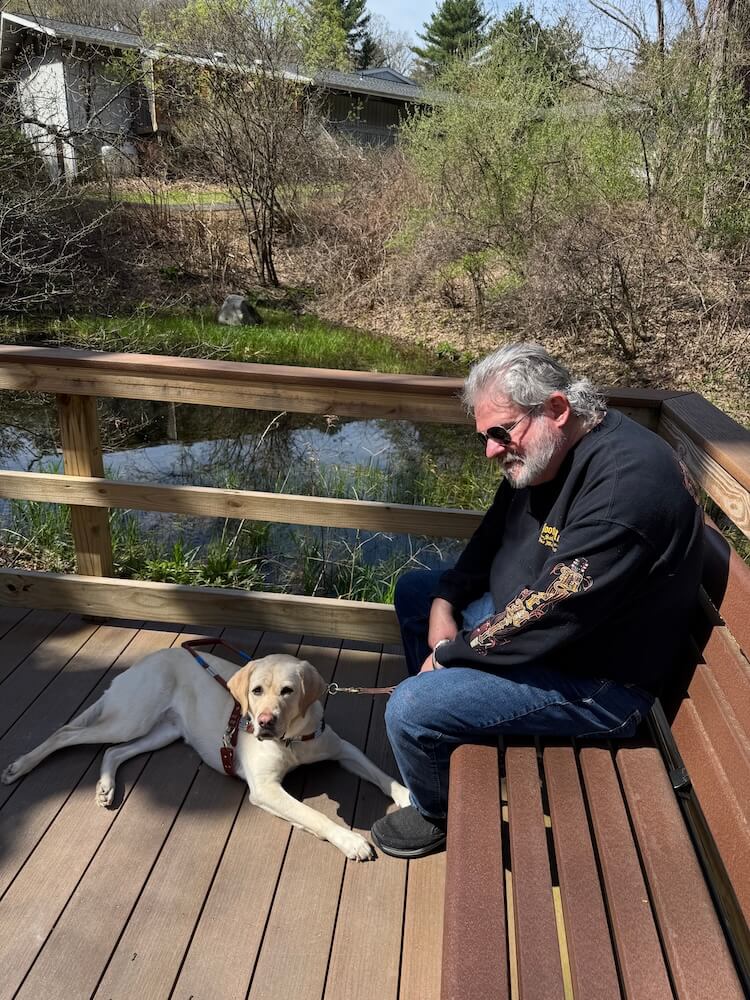 Paul sits on a wooden bench on a deck, looking down at guide Abilene lying at his feet