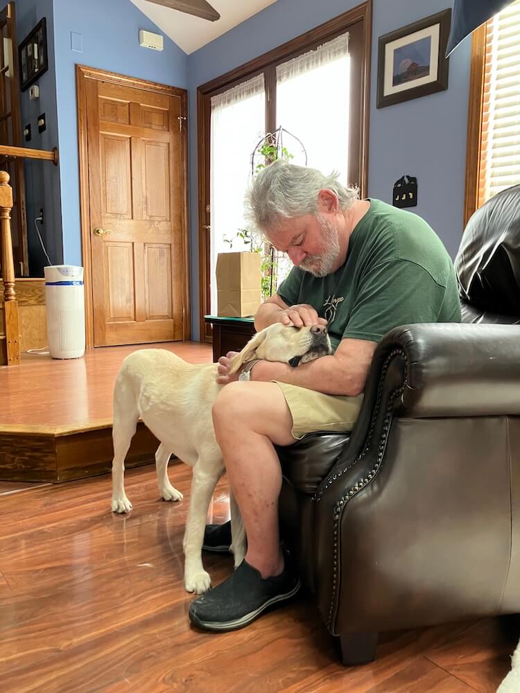 Paul sits in a chair and lovingly strokes guide Abilene's head which is contentedly on his legs with eyes closed