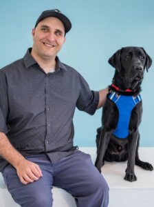 Aziz and black Lab Guide Dog Dundee pose for their graduate portrait in front of a light blue background