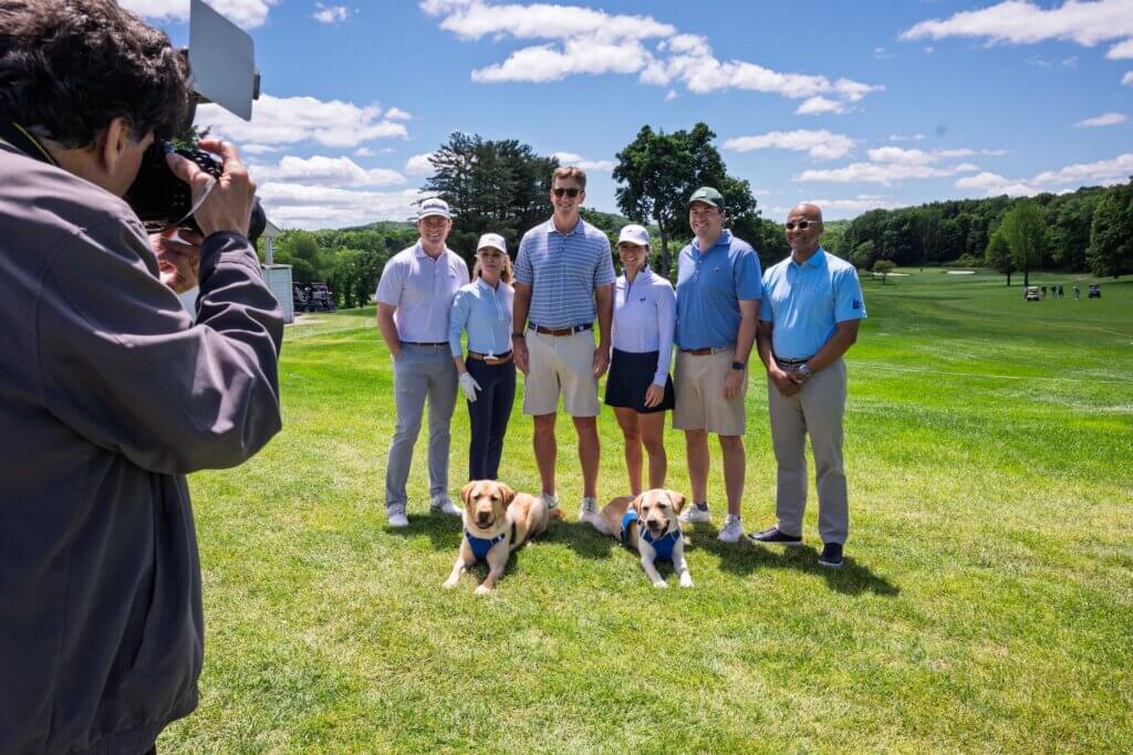 A photograher with camera in foreground shooting pic of Team of golfers with Eli Manning and Chris. Yellow guide dogs Juan (left) and Roger (right) lie on the ground