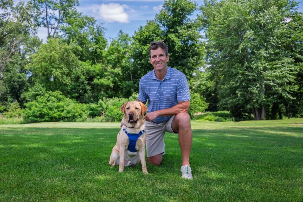 Eli on one knee on the green with yellow guide dog Juan