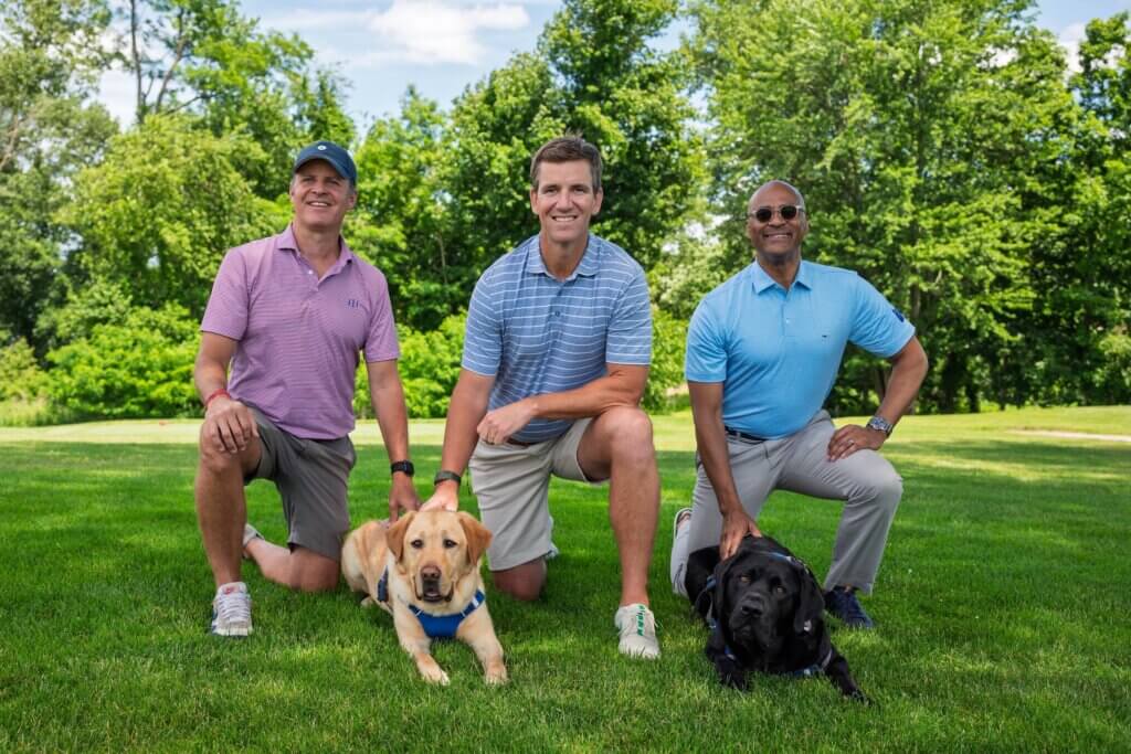 Pat Browne III, Eli, and Chris kneel with Juan (yellow) and Arnold (black), Labrador Guide dogs