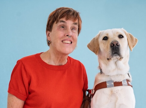 Suzanne and yellow Lab guide dog True pose for their graduate team portrait against a blue background.