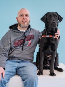 Tim and black Lab Guide Dog Omar pose for their graduate portrait in front of a light blue background