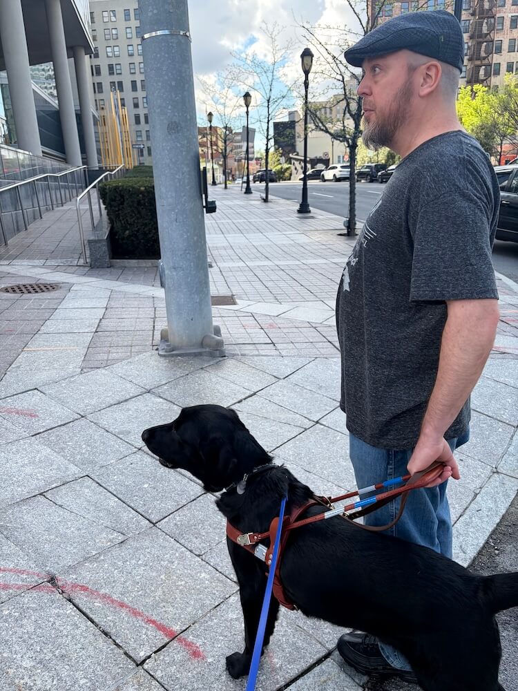 Both Tim and guide Omar look ahead as the proceed onto the sidewalk from the curb in an urban setting.