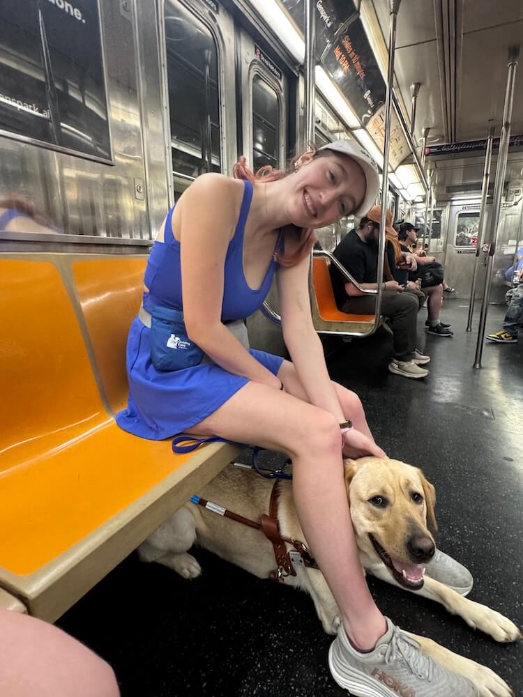 On a NYC subway car, Annalisa leans forward from a bright yellow bench to bed guide Vernon lying on the floor