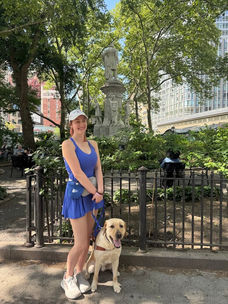 Annalisa stands with guide dog Vernon in front of a black iron fence around a statue at Verdi Square in NYC
