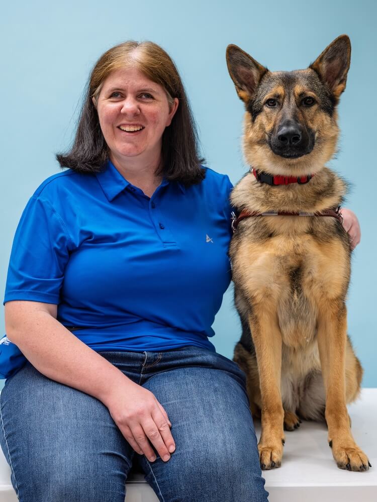 April and German Shepherd guide dog Rose sit side by side for their team portrait against a blue background