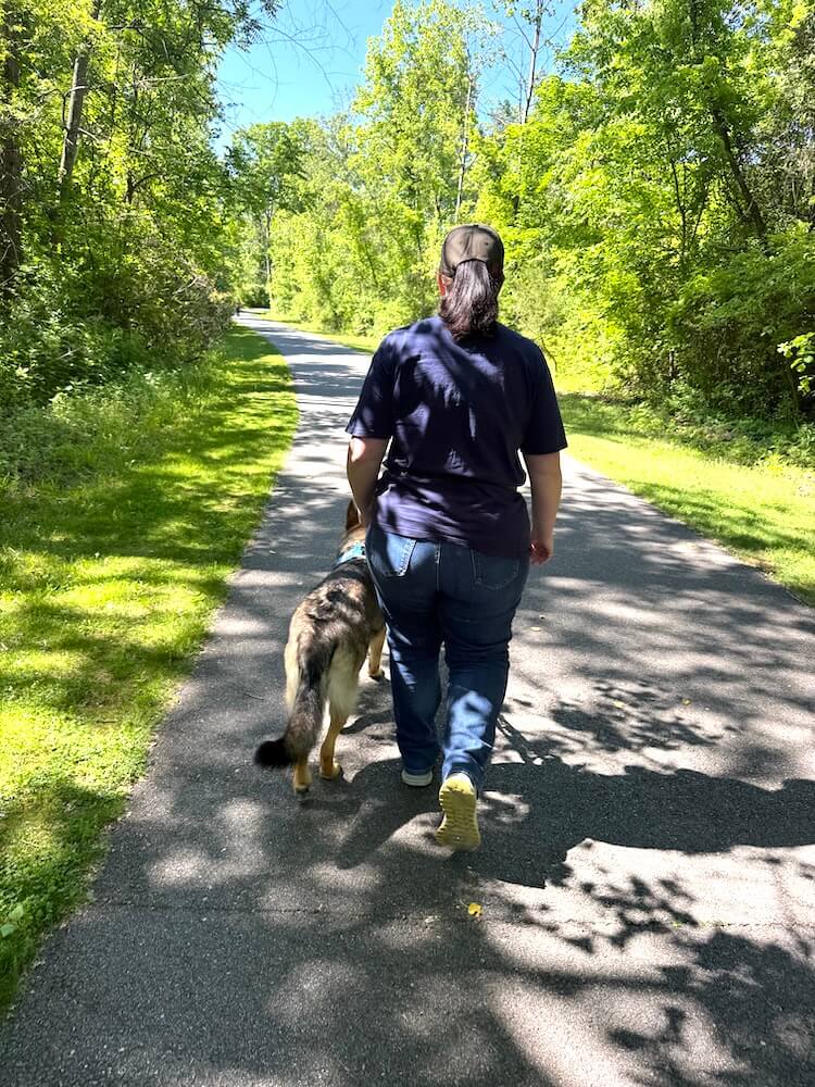 Guide dog Rose leads handler April as they walk away down a path that narrows into the trees in the distance