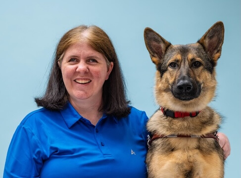 April and German Shepherd guide dog Rose sit side by side for their team portrait against a blue background