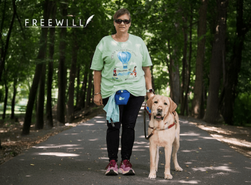 Beth and her yellow guide dog Eclipse, a male yellow Lab with a joyful expression, stand against a backdrop of a paved path through woods