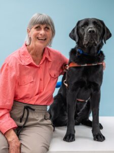 Debbie and black Lab guide dog Quidly sit side by side for their team portrait against a pale blue background