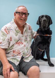 Don and black Lab guide dog Quirrel sit side by side for their team portrait against a pale blue background
