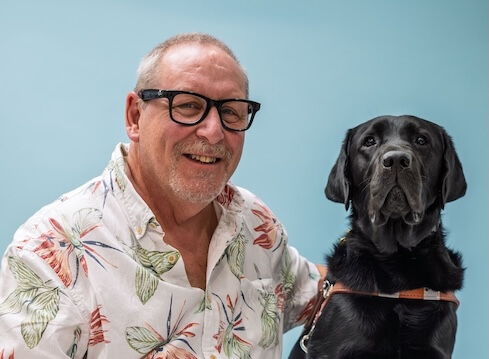 Don and black Lab guide dog Quirrel sit side by side for their team portrait against a pale blue background
