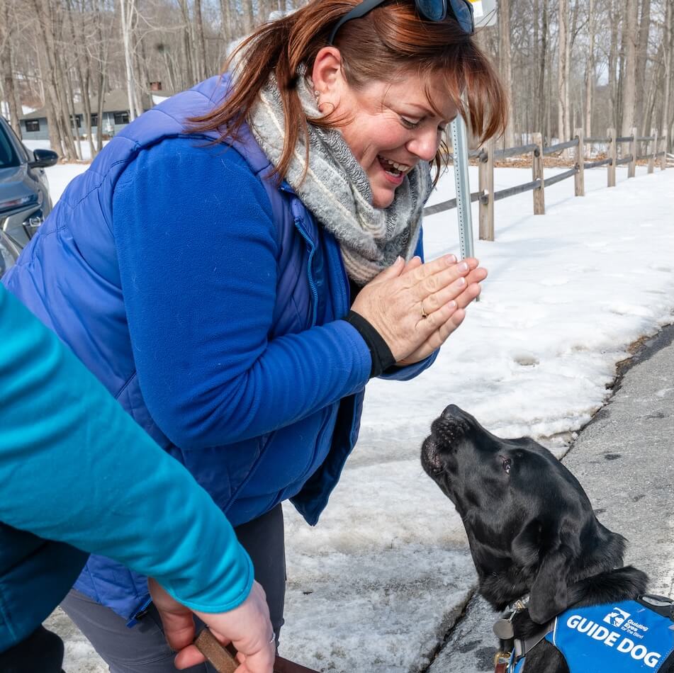 Miranda Beckmann leans forward with a happy gesture towards a black Lab guide dog in a blue harness in a snowy setting