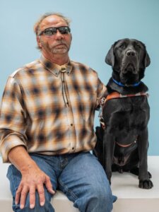 Spencer and black Lab guide dog Commander sit side by side for their team portrait against a pale blue background